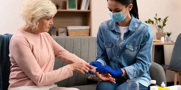 A caregiver wearing gloves and a mask checks an elderly woman's hand for health concerns.