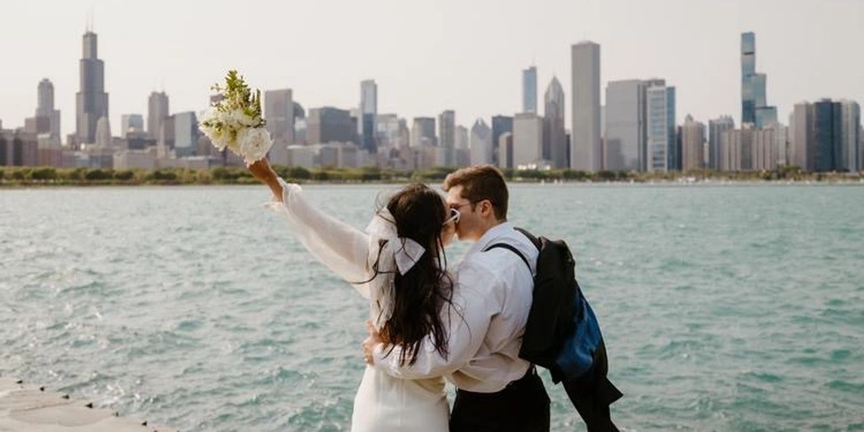 Newlywed couple kisses by the water with a city skyline in the background.