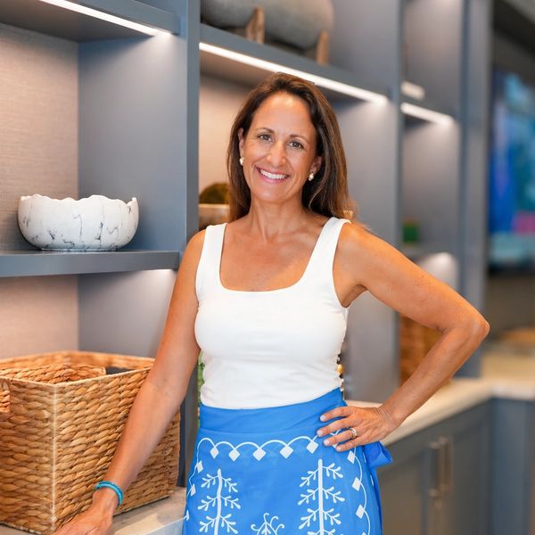 Smiling woman in a white top and blue apron stands in a modern kitchen.