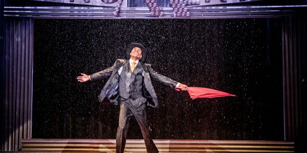 Performer joyfully dances in rain with an umbrella on stage.