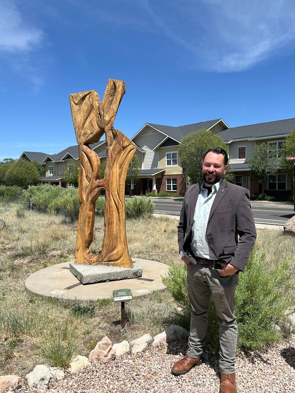 Man standing beside a tall wooden sculpture in a residential area under a clear blue sky.