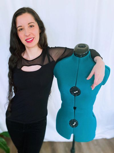 Iselda stands in front of a white backdrop with her arm around a teal green sewing dress form. 