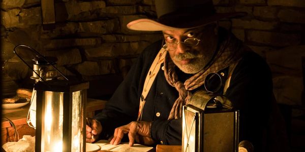 A man glances up wearing colonial gear looking at the camera. The table has two lanterns on it.