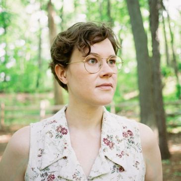 Young woman with glasses in a floral dress stands in a sunlit forest.
