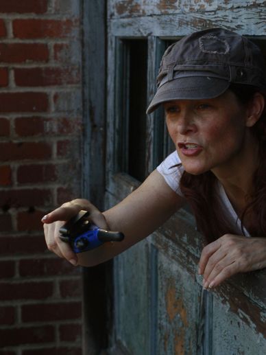 Woman in a cap pointing a blue device through a rustic window.