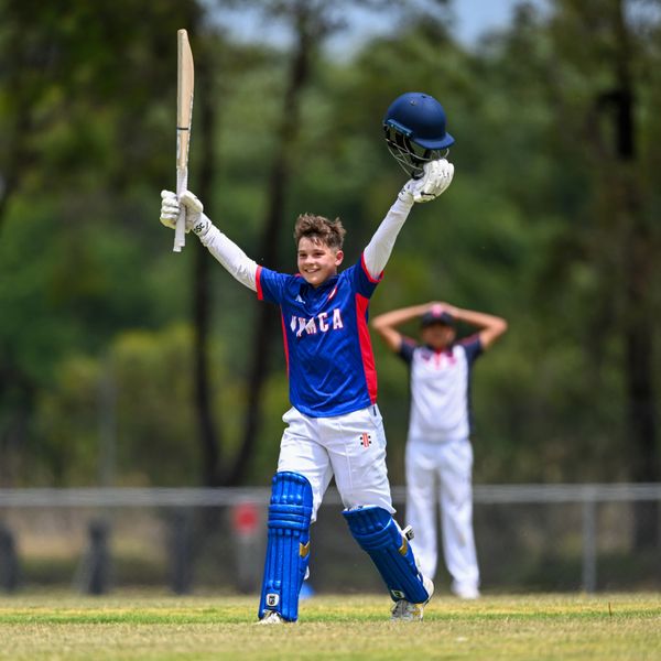 Young cricketer celebrates with bat and helmet raised on the field.