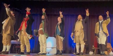 Actors in colonial costumes performing a salute on stage.