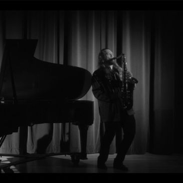A musician playing saxophone near a grand piano on stage with red curtains.