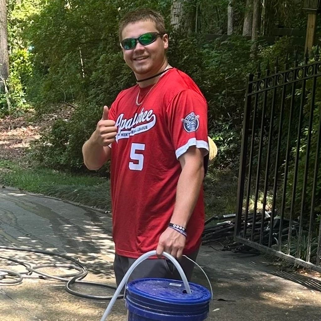 Young man in red sports shirt giving thumbs up while carrying a blue bucket outdoors.