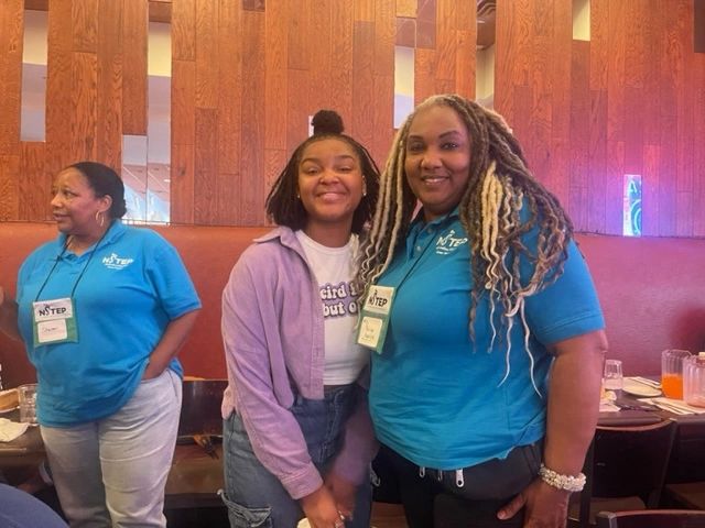 Two women smiling in a restaurant, one wearing a blue NSTEP shirt.