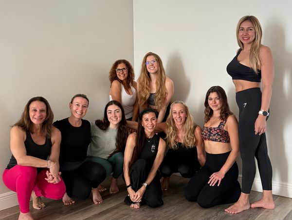 Group of nine women in workout attire posing indoors barefoot.