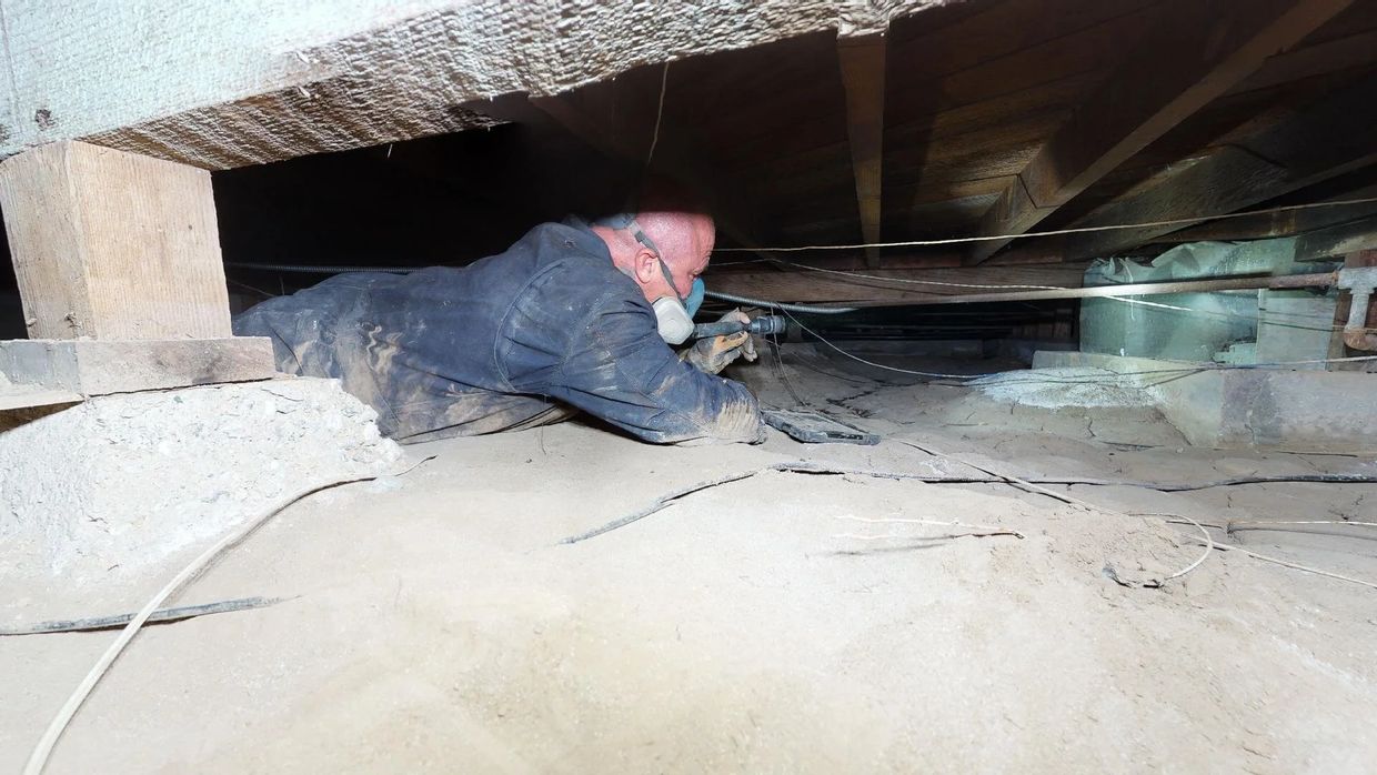 A man wearing a mask inspects a dusty crawl space under a building.
