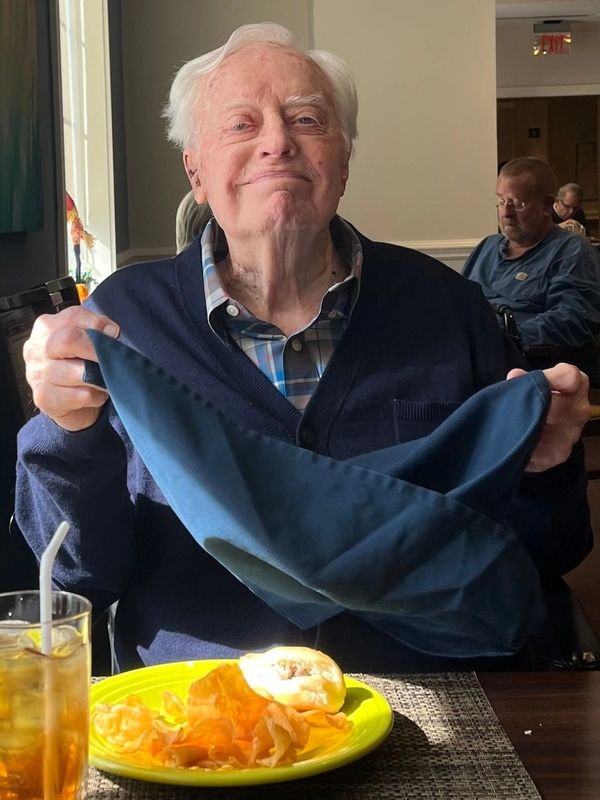 Elderly man smiling and holding a napkin at a dining table with food.