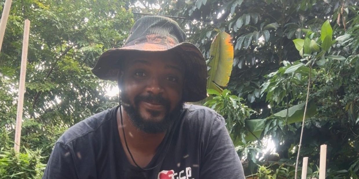 Man wearing a hat smiling outdoors surrounded by greenery.