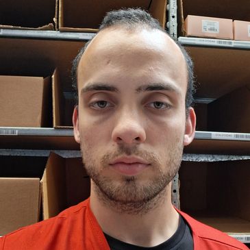 Man with short hair and beard, wearing a red shirt, standing in front of shelves with boxes.