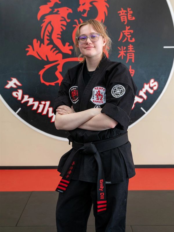 Confident martial artist in black gi with a black belt standing in front of a martial arts logo.