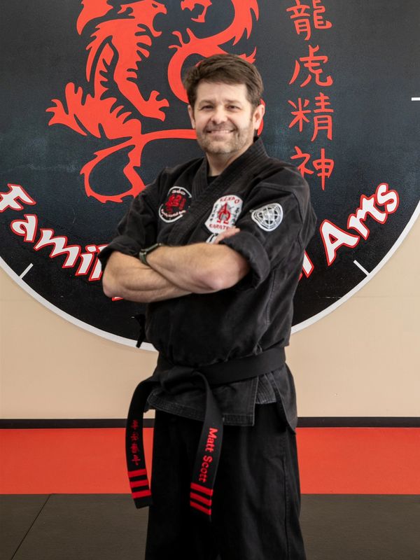 Martial artist in black gi with a black belt standing confidently in front of a martial arts logo.