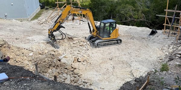 Excavator working on rocky terrain near a house under a partly cloudy sky.