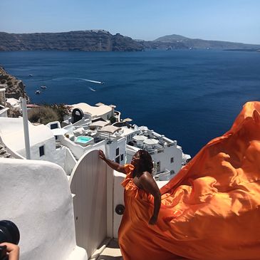 Woman in flowing orange dress overlooking a blue sea and white buildings.