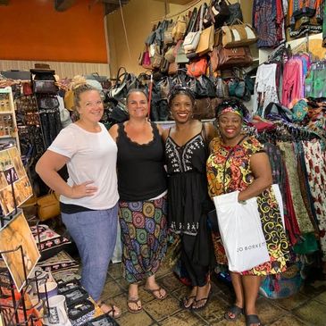 Four women smiling inside a colorful boutique filled with bags and clothes.