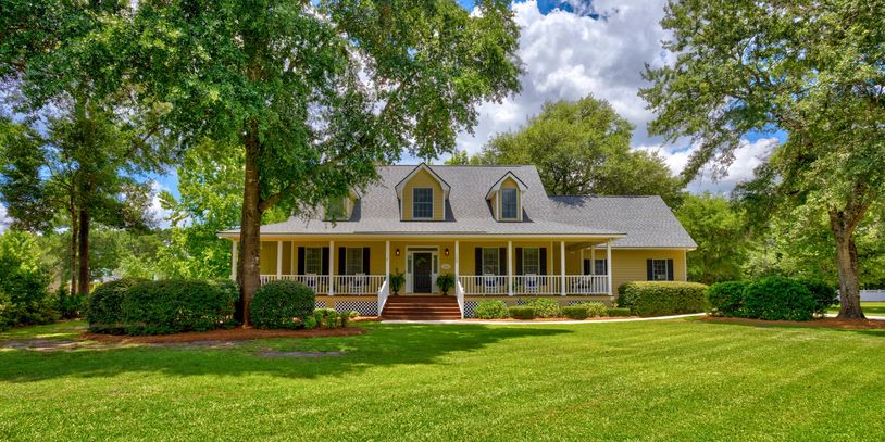 Spacious yellow house with a large porch surrounded by lush greenery and trees.