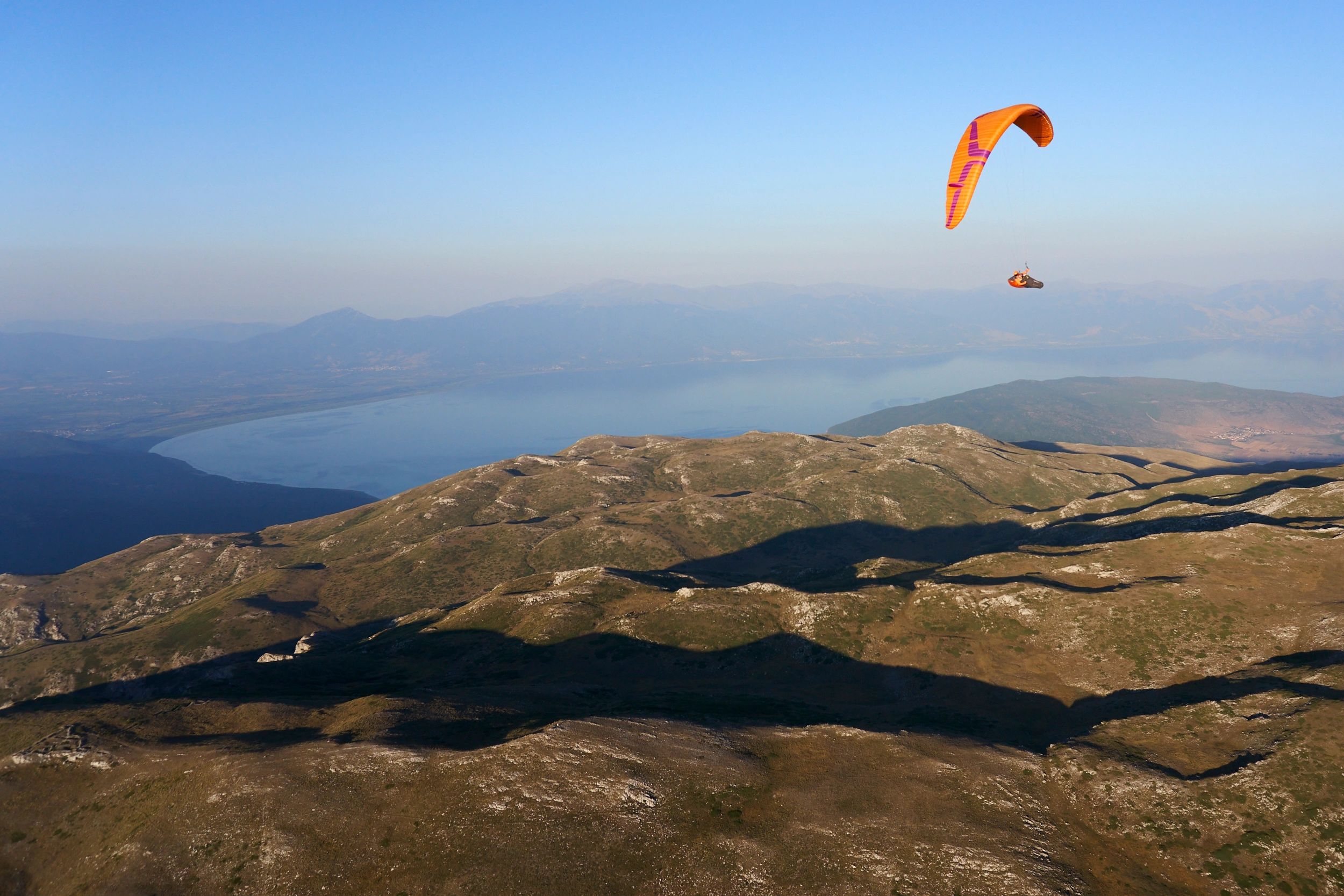 A paraglider soaring over mountainous terrain with a lake below under a clear blue sky.