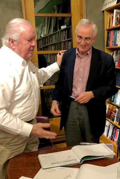 Two elderly men conversing beside a book table.