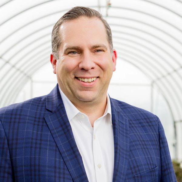 Smiling man in a blue checkered blazer inside a greenhouse tunnel.