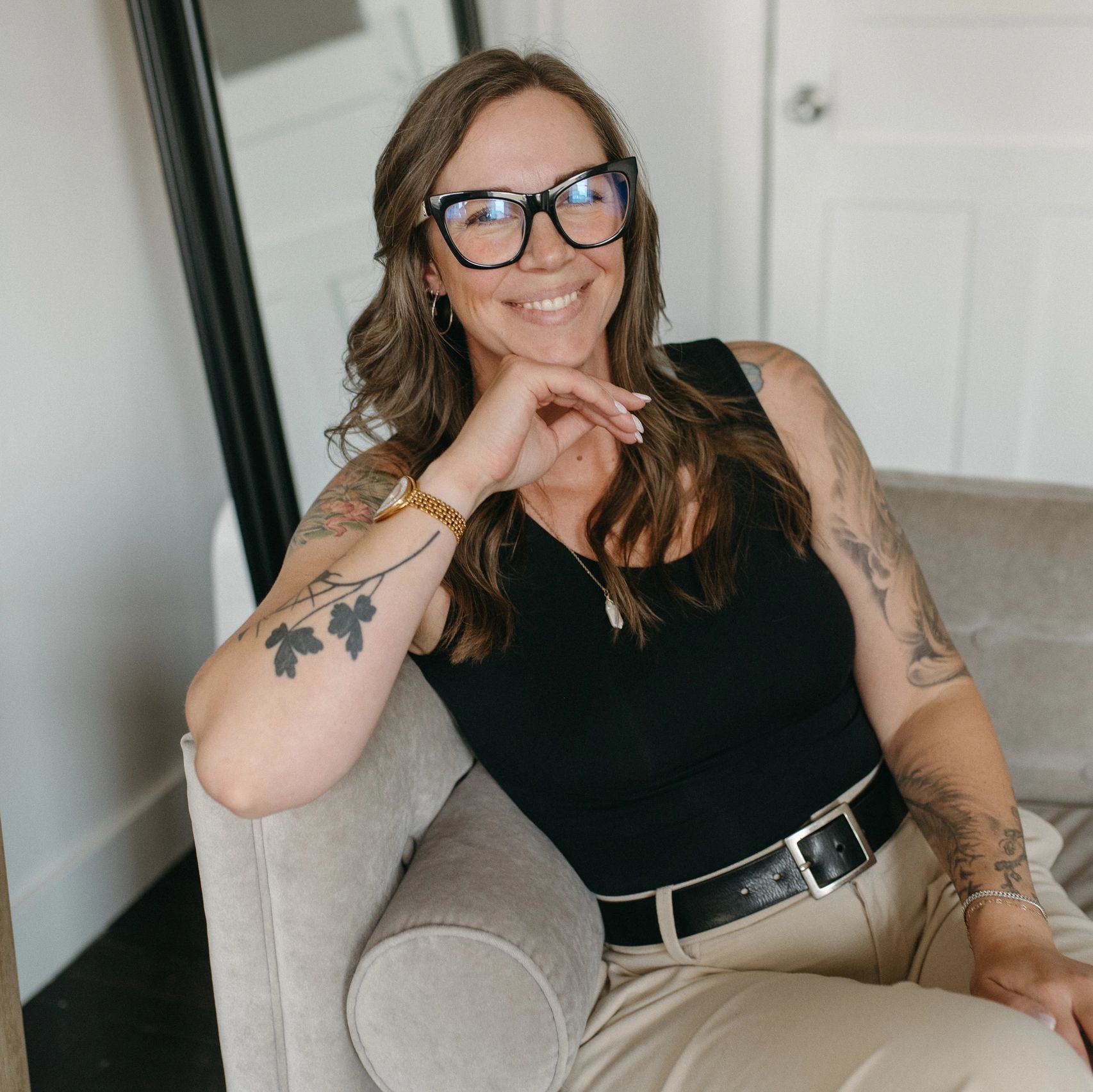 Smiling woman with glasses and tattoos sitting in a chair.