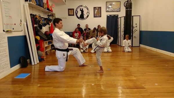A male instructor kneeling while teaching a young child how to do a kick in a dojo