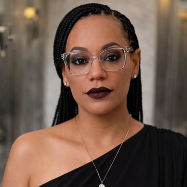 Woman with braided hair, clear glasses, and dark lipstick wearing a black one-shoulder top.