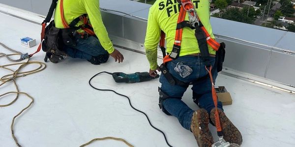 Two construction workers wearing safety harnesses working on a rooftop with a city view.