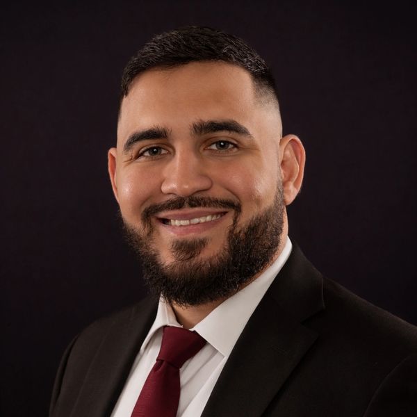 Smiling man in a suit with a red tie and beard against a dark background.
