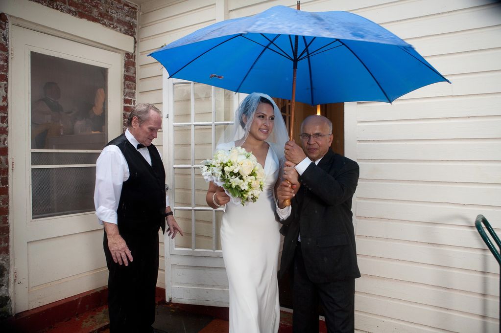 Bride and father under an umbrella while a catering waiter stands nearby in the rain