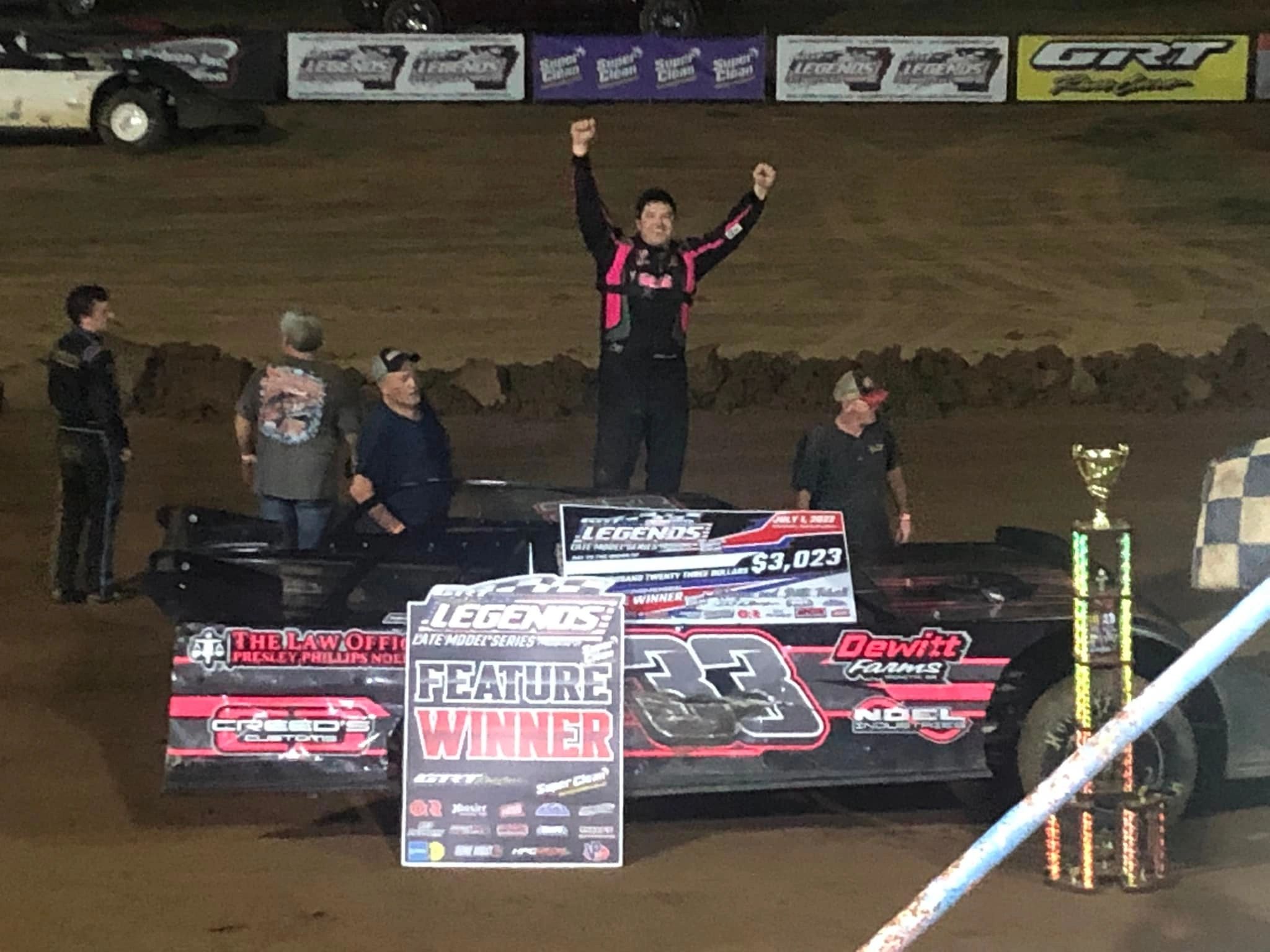 Racecar driver celebrates victory with raised arms by his car and trophy.