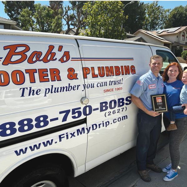 A plumber and family proudly stand next to their plumbing service van with an award plaque.