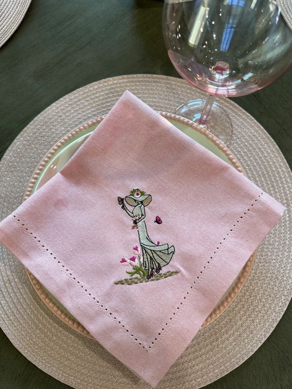 Pink embroidered napkin on a decorative plate with a wine glass.