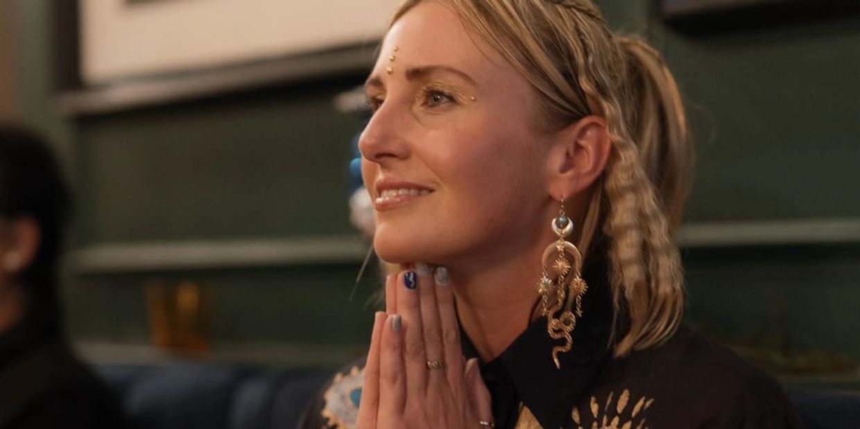A woman with intricate earrings and face adornments smiles thoughtfully.