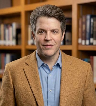 Man in a brown blazer and blue shirt in a library setting.