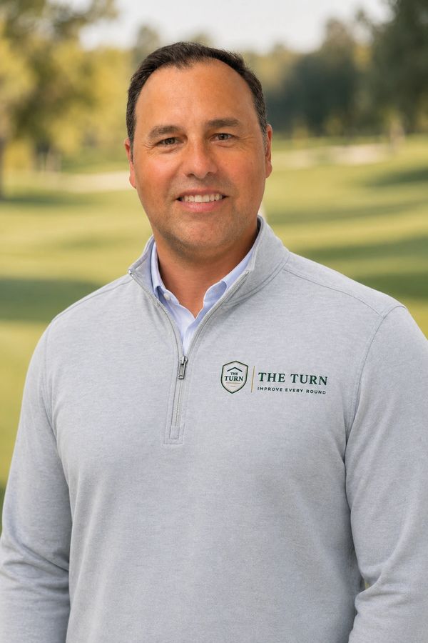 Smiling man wearing a light gray zip-up sweater with 'The Turn' logo outdoors.