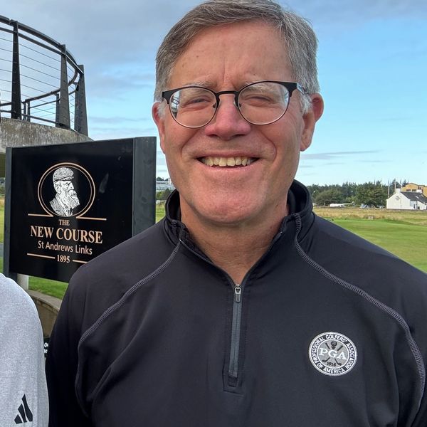 Smiling man in glasses wearing a PGA jacket at St Andrews golf course.
