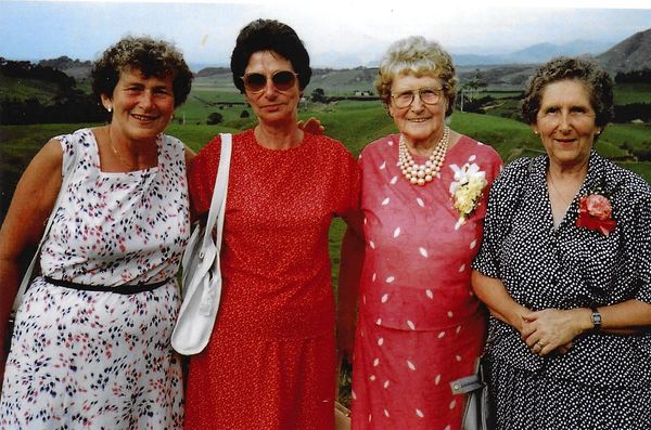 Four elderly women posing outdoors with countryside in the background.