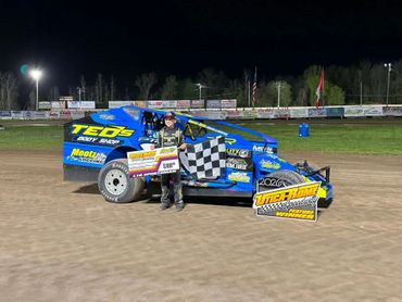 Race car driver posing with a checkered flag beside a blue race car at night.