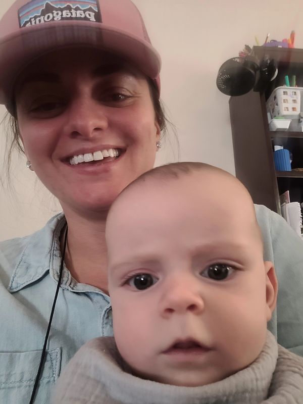 A smiling woman wearing a pink hat holds a curious baby close to the camera.