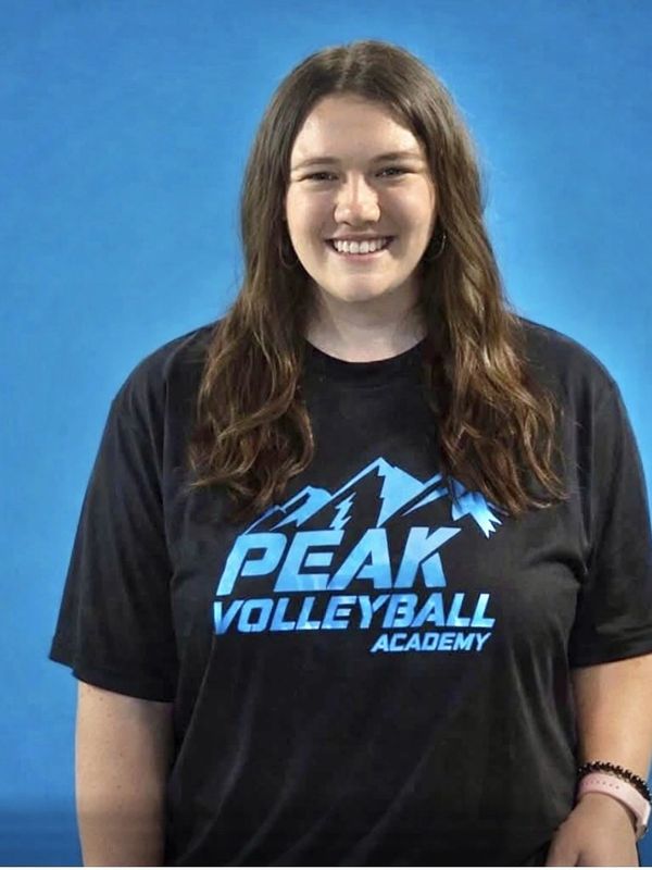 Young woman smiling in a Peak Volleyball Academy t-shirt against a blue background.