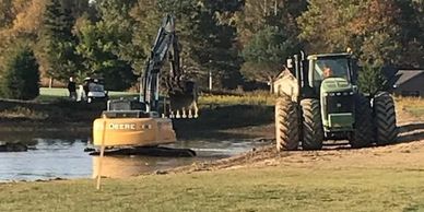 Excavator and tractor near a pond in a grassy area.