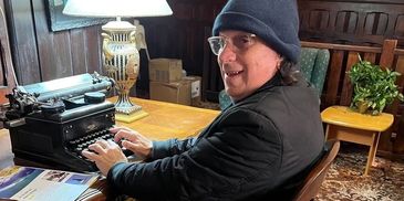 Man in a hat typing on a vintage typewriter at a wooden desk.