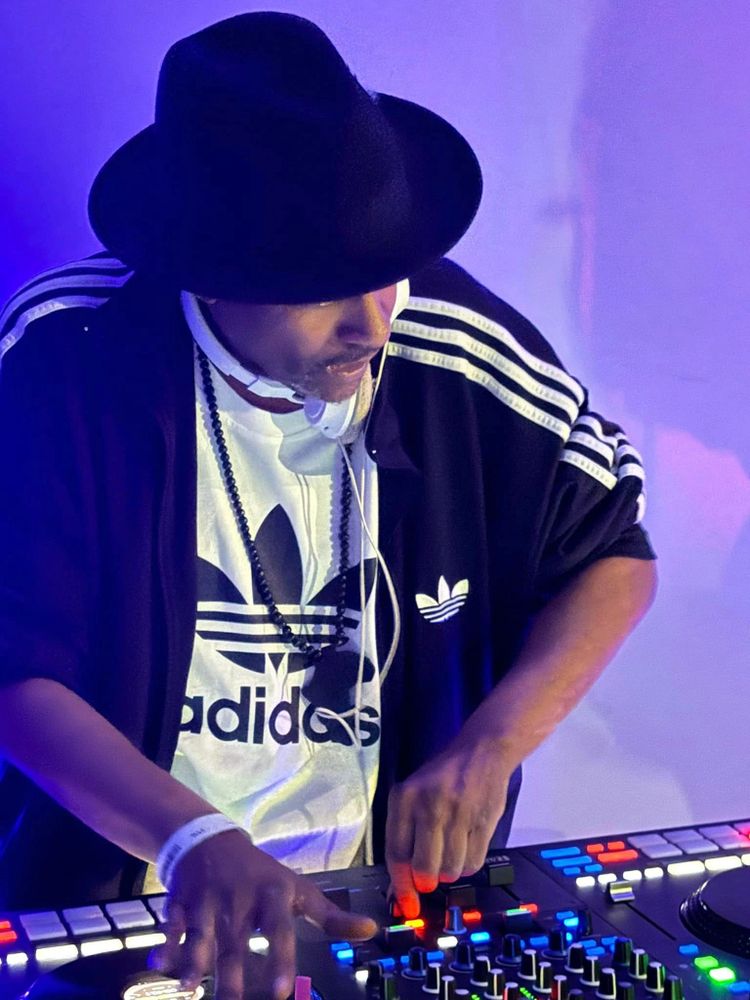 DJ wearing a black hat and Adidas jacket performing with a mixer.