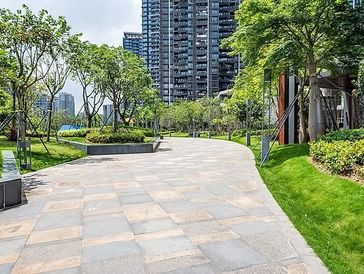 A paved pathway winds through a green urban park with tall buildings in the background.