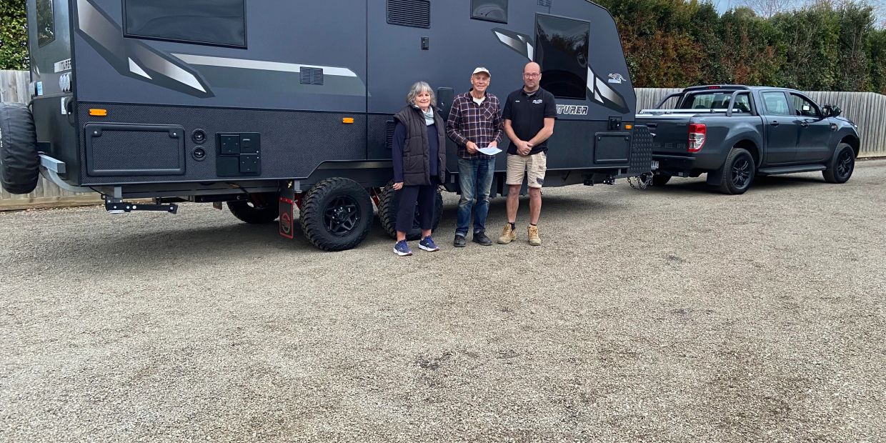 Three people standing in front of a large black camper trailer hitched to a pickup truck.